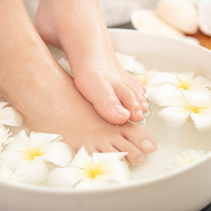 closeup view of woman soaking her feet in dish with water and fl closeup view of woman soaking her feet in dish with water and flowers on wooden floor. Spa treatment and product for female feet and hand spa. white flowers in ceramic bowl.
